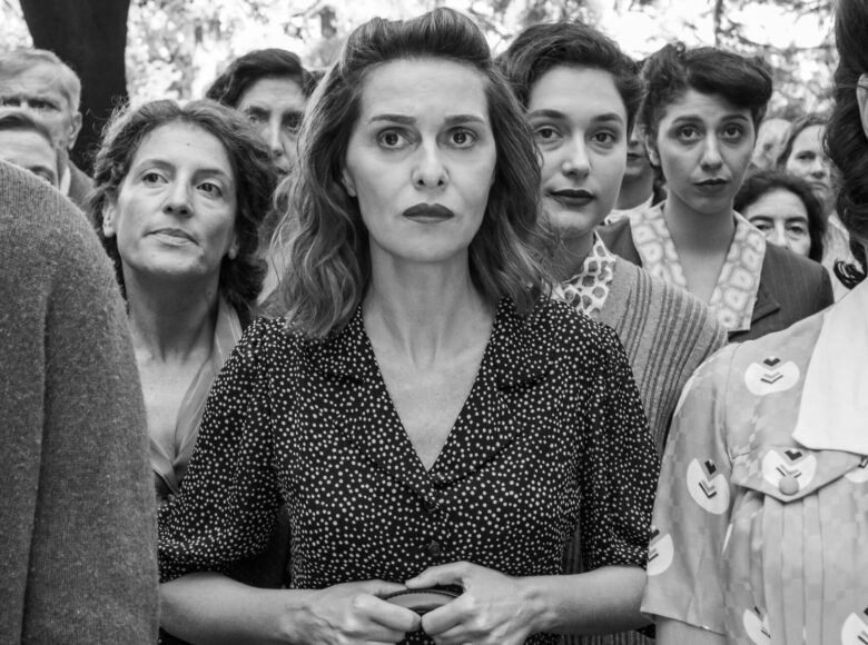 Czarno-biała fotografia kilku kobiet zgromadzonych we wspólnej sprawie. Wyrazy twarzy kobiet są różne, słuchają i wpatrują się we wspólny punkt.
