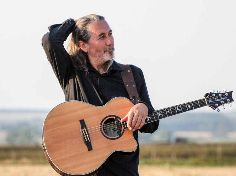 Kolorowe zdjęcie. Przedstawia mężczyznę, który stoi na środku pole. Na ramieniu ma zawieszona gitarę akustyczną.
