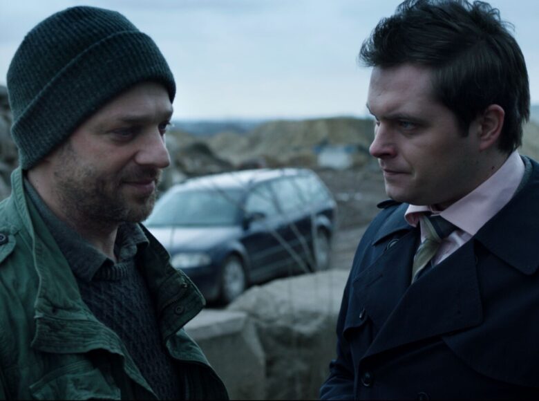 Kadr z filmu. Na zewnątrz. Stoi dwóch mężczyzn, jeden z zarostem, w swetrze, zielonej kurtce, czapce, drugi ogolony, pod krawatem.