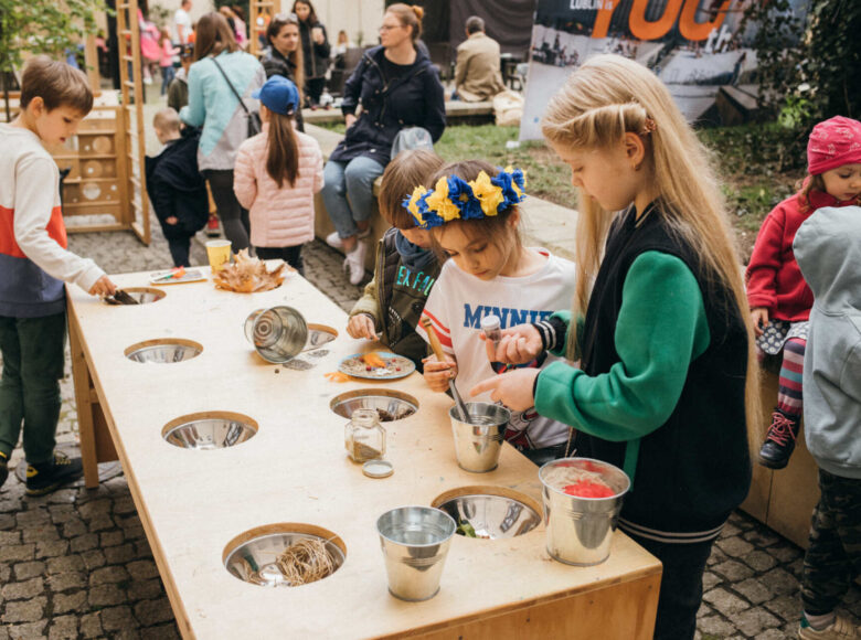 Zdjęcie przedstawia małą grupę dzieci, która bawi się przy drewnianym stole. Na stole znajdują się miski z koralikami, piaskiem i sznurkami.