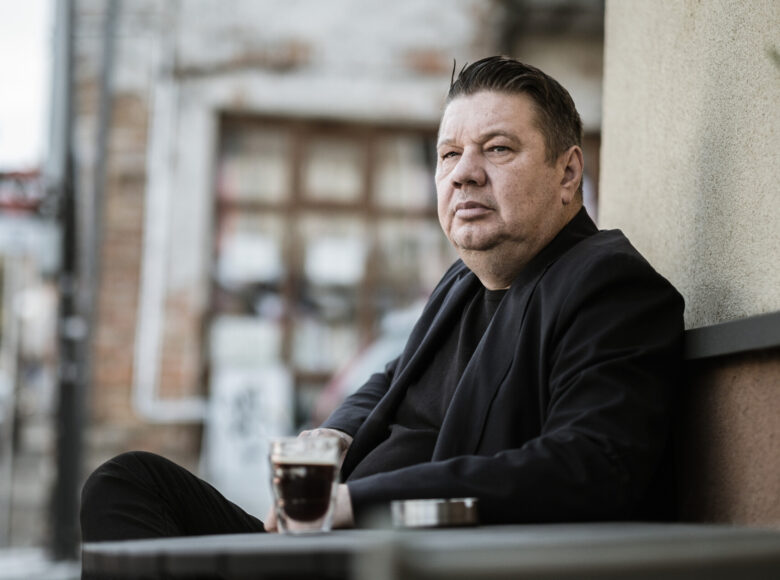 Na zdjęciu mężczyzna siedzący przy stoliku w kawiarni. Oparty plecami o ścianę budynku. Jest ubrany w czarną koszulkę i czarną marynarkę. Patrzy przed siebie. Na stole mała kawa espresso i popielniczka.