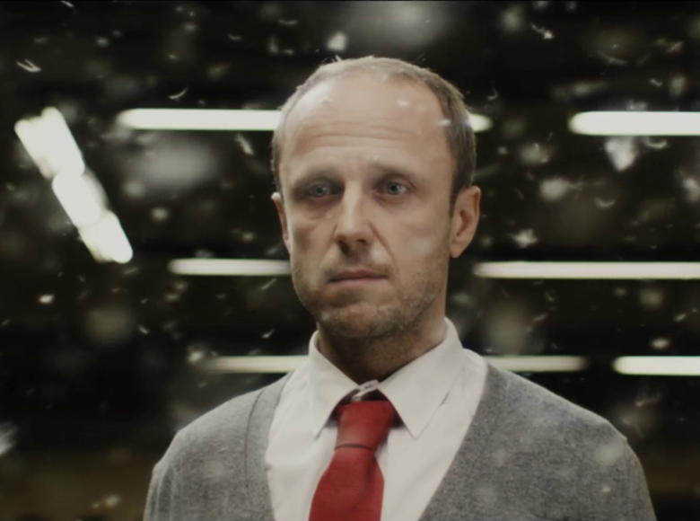 Kadr z filmu. Wnętrze. Mężczyzna w szarym sweterku, białej koszuli i czerwonym krawacie patrzy przed siebie. W tle świetlówki, w powietrzu unosi się pierze.