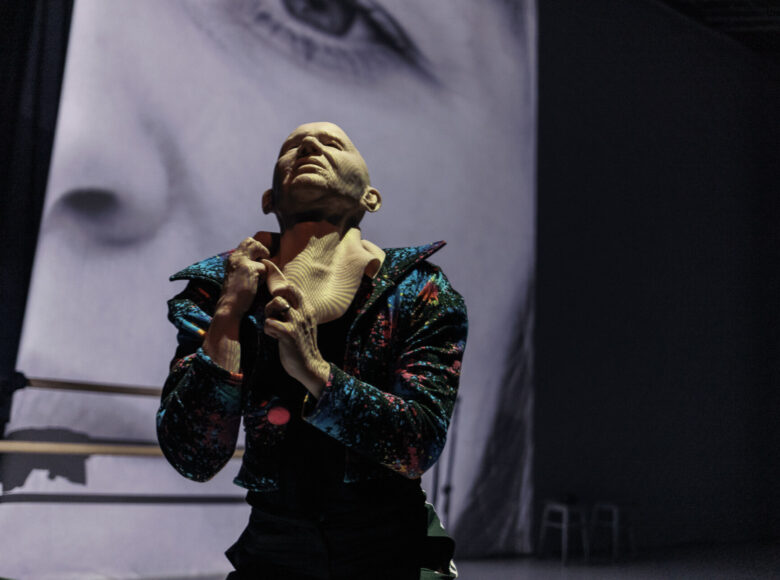 Zdjęcie ze spektaklu teatru Tańca. Na scenie znajduje się osoba w kolorowym kostiumie, która zdziera z siebie maskę.