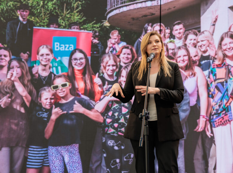 Do mikrofonu przemawia zastępczyni prezydenta Lublina do spraw kultury. Za nią na ścianie wielkoformatowa fotografia, przedstawiająca grupę młodych ludzi.