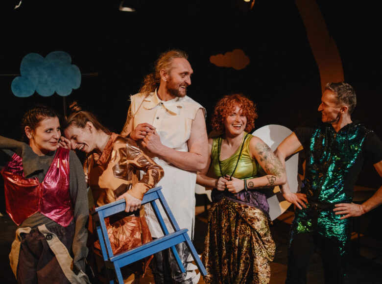 Spektakl teatralny. Grupa roześmianych aktorek i aktorów w barwnych, błyszczących strojach stoi jedno za drugim. W tle dekoracje przedstawiające kolorowe chmurki na czarnym tle.