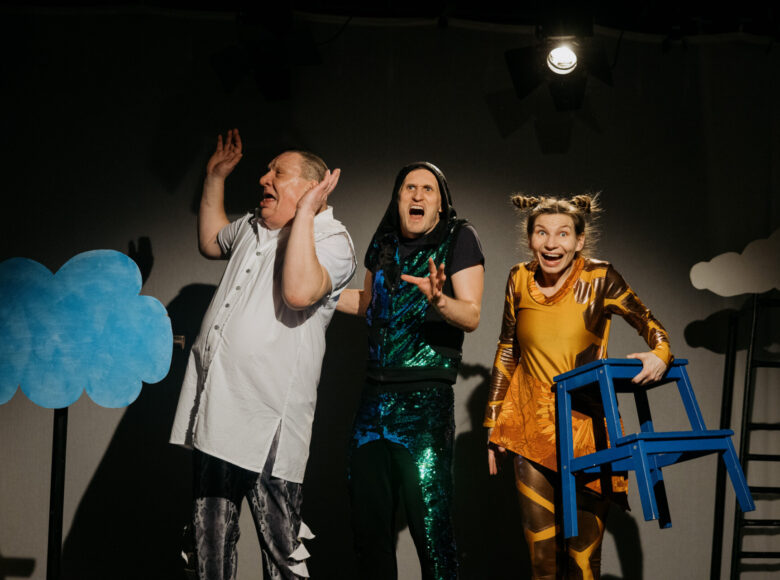Spektakl teatralny. Aktorka i dwóch aktorów stoją w rzędzie obok siebie, przybierając dynamiczne pozy. Ubrani są w bajkowe kostiumy. Wokół nich dekoracje przedstawiające chmurki.