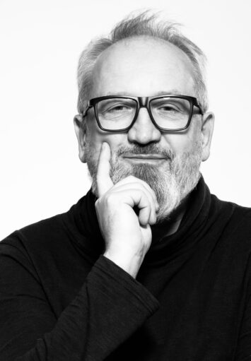 Zdjęcie przedstawia portret mężczyzny ubranego w czarny golf. Mężczyzna nosi okulary, placem wskazującym dotyka swojej brody. Tło białe, jednolite.