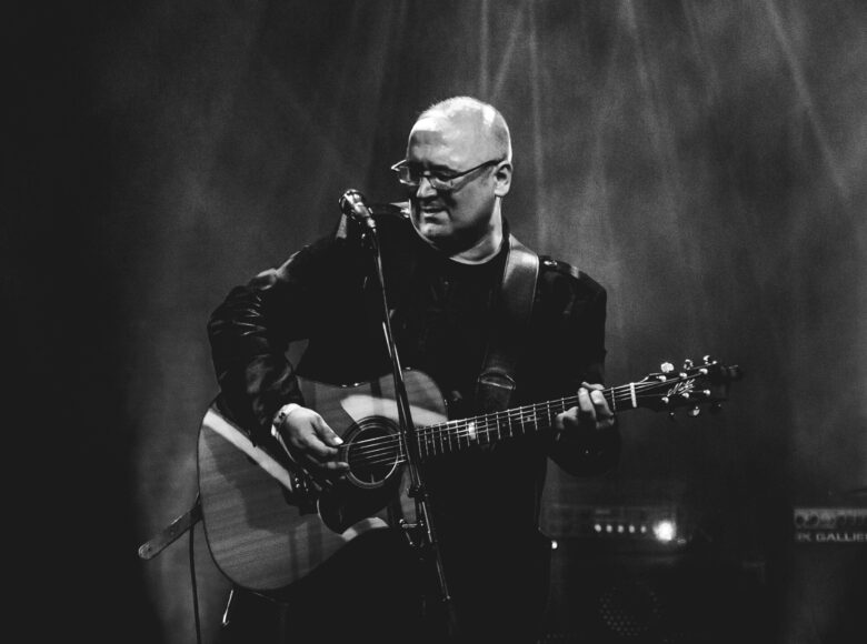 Czarno-białe zdjęcie. Przedstawia mężczyznę, który znajduje się na scenie i gra na gitarze. Padają na niego światła z reflektorów.