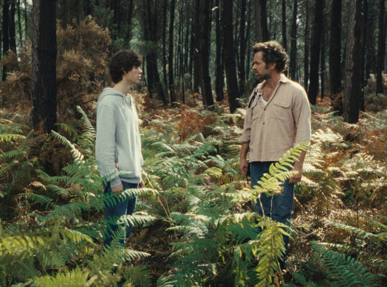 Fotos z filmu przedstawia młodego chłopaka i trochę starszego mężczyznę stojących wśród drzew i paproci w lesie. Chłopak ubrany jest w szarą bluzę i niebieskie dżinsy, a mężczyzna w brązową koszulę i niebieskie dżinsy.