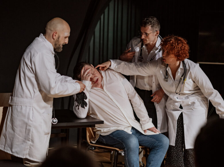 Fotografia przedstawia aktorów, przebranych za lekarzy. W centrum siedzi główny bohater. Po jego lewej stoi mężczyzna w fartuchu nakładający rękawiczki, po prawej mężczyzna przyglądający się sytuacji, oraz kobieta dotykająca palcem głównego bohatera.