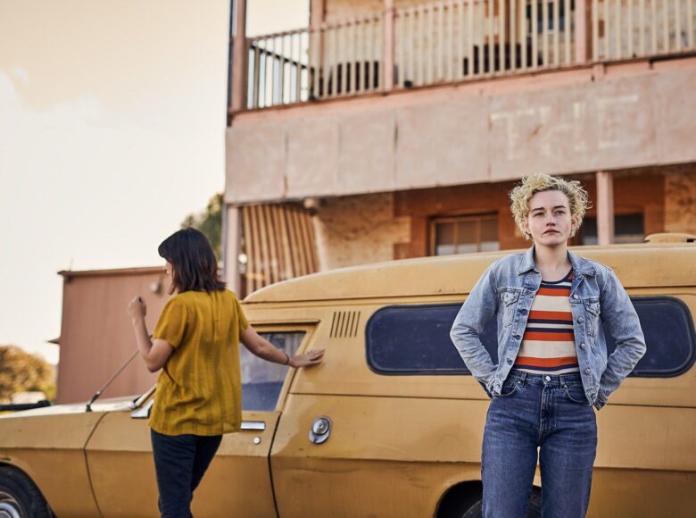 Kolorowa fotografia przedstawia dwie kobiety na tle auta i budynku. Kobieta po lewej, w żółtej bluzce stoi tyłem do obiektywu. Druga stoi po prawej, ubrana jest w niebieskie dżinsy i dżinsową kurtkę oraz bluzkę w paski. Patrzy się w dal.