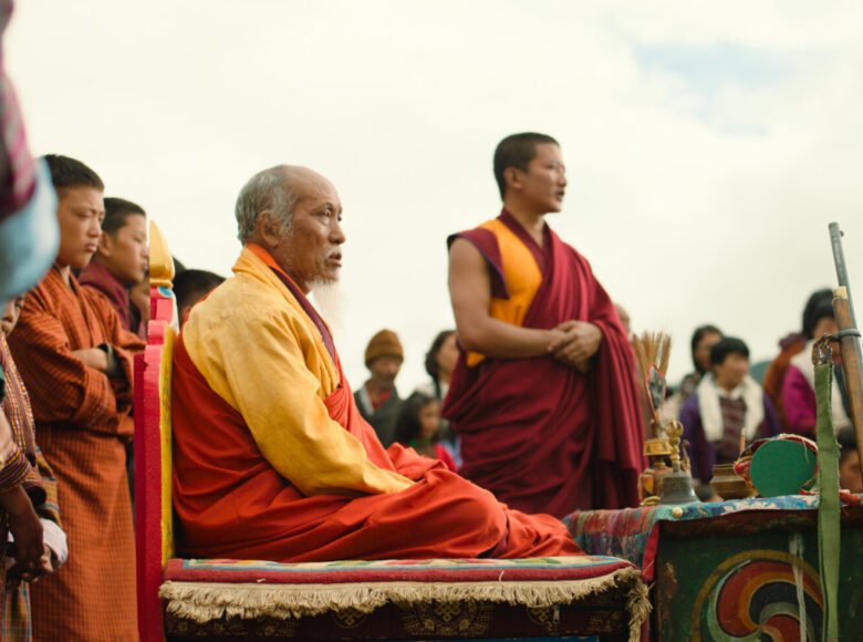 Kolorowa fotografia przedstawia zgromadzone osoby ubrane w tradycyjne stroje mnichów. Główna postać na zdjęciu to siedzący, starszy mężczyzna. Wszyscy patrzą w tą samą stronę.