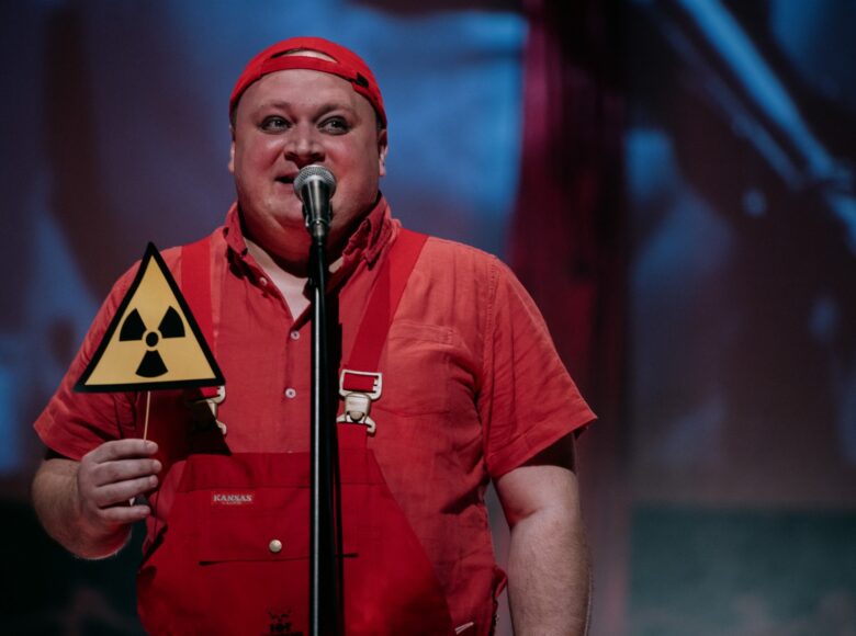 Aktor ubrany w czerwoną koszulę i czerwony kombinezon trzyma w ręku znak radiacji na patyku przypominający lizak. Śpiewa do mikrofonu, uśmiechając się. Tło jest rozmazane.