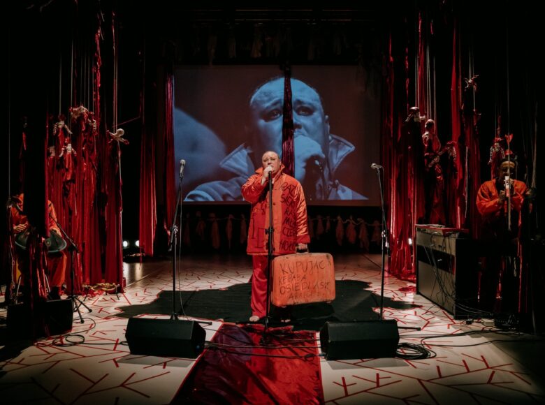 Pośrodku sceny stoi aktor śpiewający do mikrofonu. W tle znajduje się czarno-biała projekcja z jego zbliżeniem na twarz. Jest ubrany na czerwono, jest bosy i trzyma w ręku czerwoną walizkę z napisem "KUPUJĄC KEBABA OSIEDLASZ ARABA". Po obu stronach sceny, nieco schowani za długimi czerwonymi lalkami motankami zwisającymi z sufitu, znajdują się dwaj muzycy. Podłoga jest biała, a pośrodku leży czarny dywan w kształcie mapy Białorusi, na którym stoi aktor.