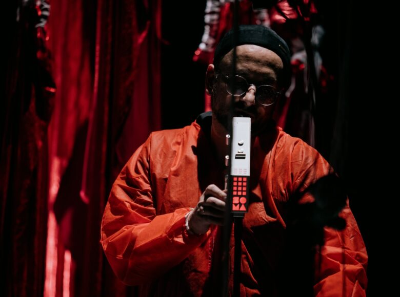 Muzyk w czerwonym kombinezonie i okularach gra na instrumencie. Tło jest rozmazane, a dominują kolory czerwony i czarny.