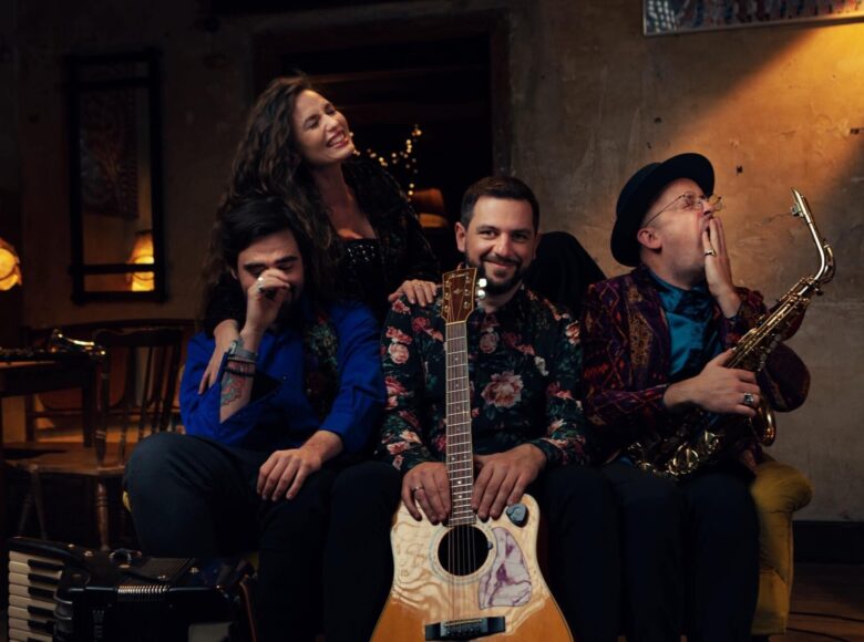 Kolorowa fotografia przedstawia czterech muzyków o dobrych humorach. Od lewej mężczyzna w niebieskiej koszuli, w środku siedzi mężczyzna w koszuli w kwiaty z gitarą, a po prawej siedzi mężczyzna w kolorowej marynarce z saksofonem. Nad nimi stoi śmiejąca się kobieta o długich, ciemnych włosach, ubrana na czarno.