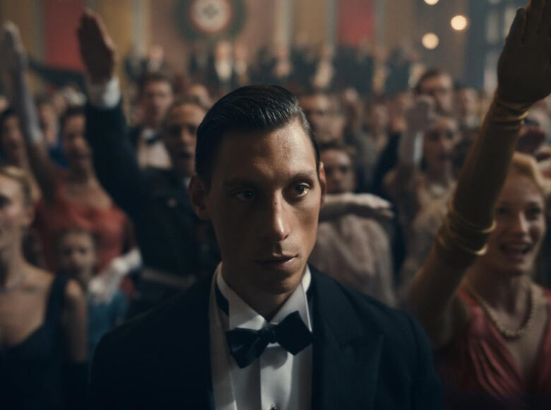 Kolorowy fotos z filmu "Filip". Na pierwszym planie w centrum zdjęcia znajduje się tytułowy bohater w garniturze. Mężczyzna ma pociągłą twarz, ciemne oczy i włosy zaczesane do tyłu. W tle bohatera stoi wiele osób ubranych na galowo.