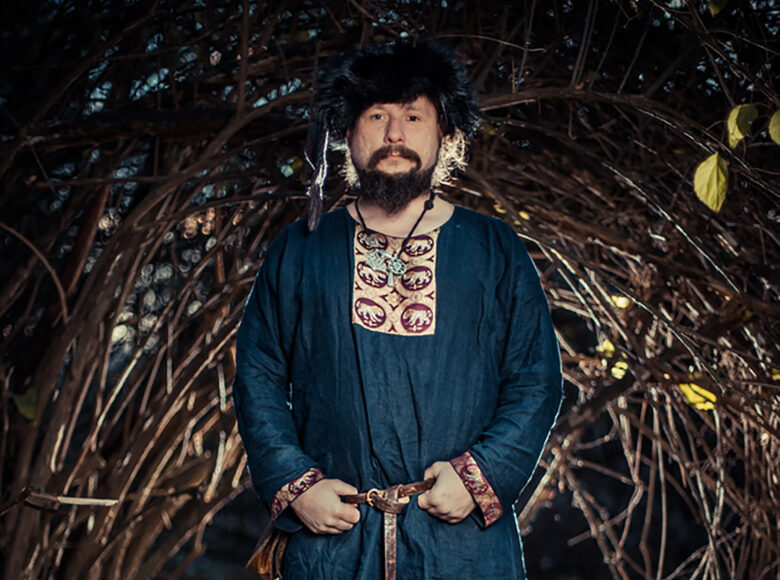 Kolorowa, ale ciemna fotografia przedstawia mężczyznę starym, ciemnoniebieskim, średniowiecznym ubiorze. Mężczyzna stoi w centrum fotografii, wśród wielu gałęzi.