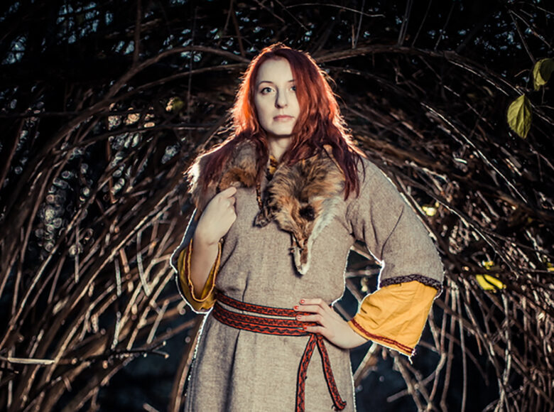 Kolorowa, ale ciemna fotografia przedstawia kobietę starym, jasnobrązowym, średniowiecznym ubiorze. Kobieta stoi w centrum fotografii, wśród wielu gałęzi.