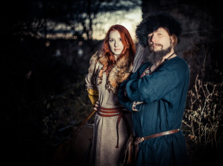 Kolorowa, ale ciemna fotografia przedstawia kobietę i mężczyznę w starych, średniowiecznych ubraniach stojących w lesie. Kobieta ubrana jest na jasnobrązowo, a mężczyzna na ciemnoniebiesko. Oboje skierowani są w stronę obiektywu.
