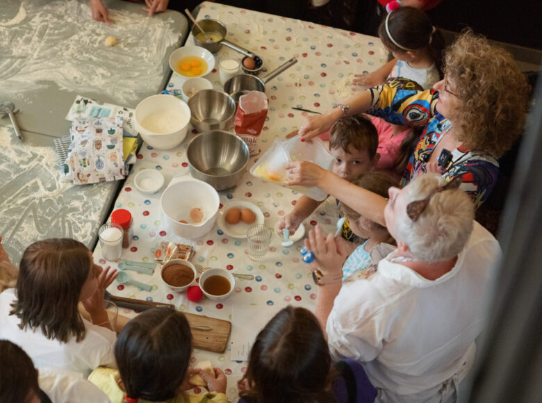 Kolorowe zdjęcie przedstawia stół ze składnikami i naczyniami do pieczenia, a wokół niego zebrane są osoby w różnym wieku.