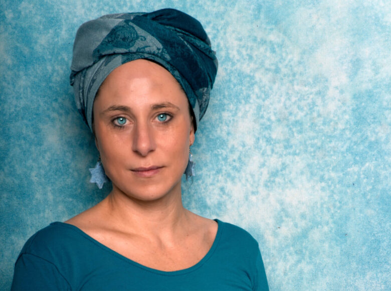 Zdjęcie przedstawia popiersie kobiety o niebieskich oczach, w niebieskiej bluzce oraz niebieskiej chuście na głowie. Kobieta stoi bliżej lewej krawędzi zdjęcia na niebieskim tle.