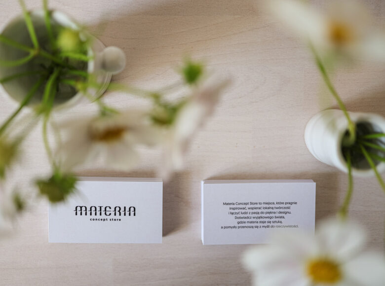 Kolorowe zdjęcie przedstawia rozmazane, zielone kwiaty w dwóch rogach kadru. Natomiast obiektyw skupia się na dwóch, białych kartkach. Po lewej kartka z logiem "MATERIA concept store", ta po prawej opisuje czym jest Materia Concept Store".