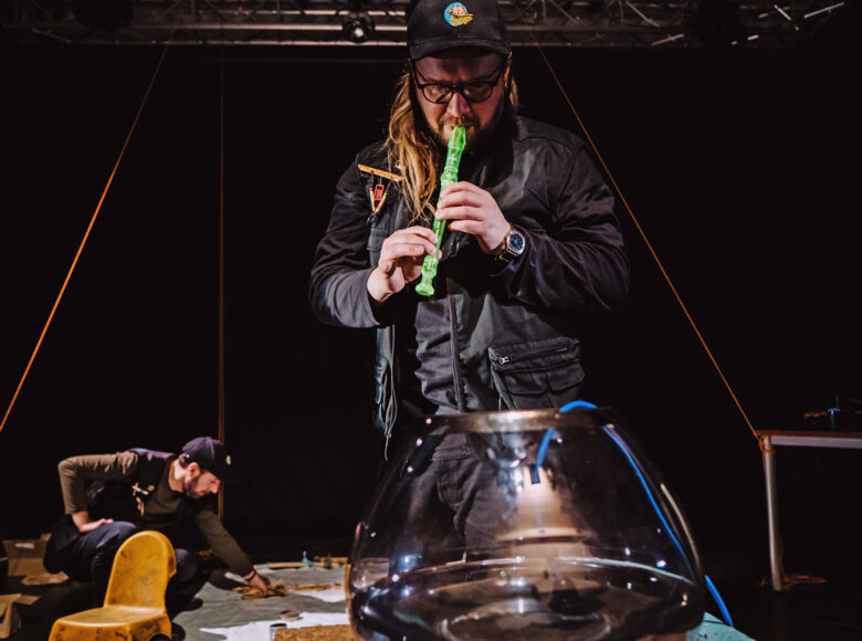 Na zdjęciu dwóch mężczyzn. Na pierwszym planie półprzezroczyste naczynie z wężem oświetlonym na niebiesko. Za nim stoi mężczyzna grający na flecie prostym, w kolorze jaskrawo seledynowym. W tle klęczy drugi mężczyzna, rozkładający przedmioty na pasie jasnego materiału.