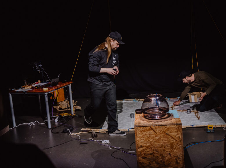 Na zdjęciu dwóch mężczyzn w przestrzeni sceny. Na pierwszym planie postument ze sklejki, na którym stoi szklana kula-akwarium. Za postumentem rozłożona na ziemi biała tkanina. Mężczyzna po prawej klęczy, rozkładając przedmioty na tkaninie. Drugi idzie po lewej stronie materiału. Po lewej stronie kadru stół z instrumentami elektronicznymi.