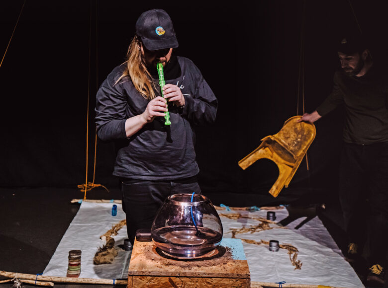 Na zdjęciu dwóch mężczyzn. Na pierwszym planie półprzezroczyste naczynie z wężem oświetlonym na niebiesko. Za nim stoi mężczyzna grający na flecie prostym, w kolorze jaskrawo seledynowym. W tle w tle trzyma niskie plastikowe krzesło, przenosi je nad jasnym materiałem rozłożonym na podłodze.