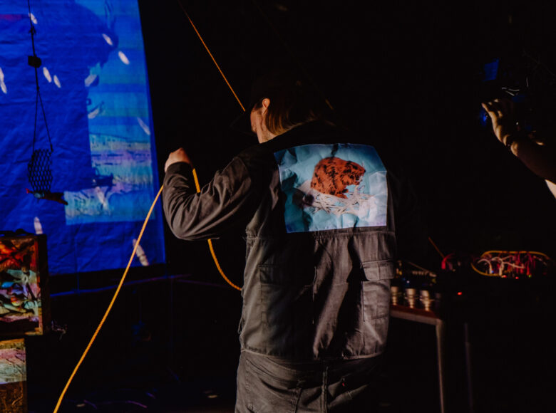 Mężczyzna stojący w centrum kadru, odwrócony tyłem. Na plecach kurtki ma grafikę przedstawiającą bobra na niebieskim tle. W rękach trzyma oświetlone na pomarańczowo sznurki. W tle oświetlony na niebiesko ekran, przed którym stoją oświetlone na pomarańczowo postumenty.