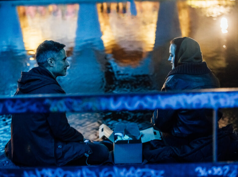 Zdjęcie o niebieskich barwach przedstawia dwójkę osób, mężczyznę i kobietę siedzących nad wodą. Oboje są zwróceni do siebie, a między nimi stoi otwarte pudełko.