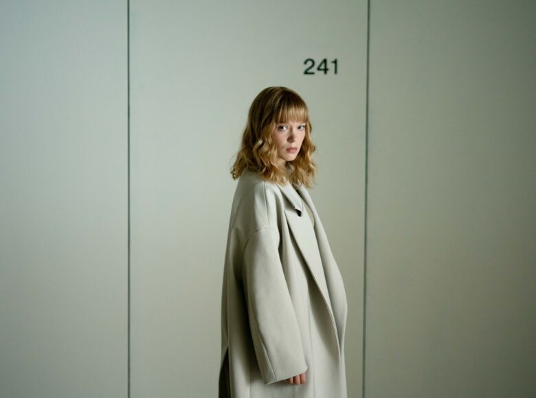 Jasna, kolorowa fotografia przedstawia kobietę w centrum zdjęcia. Ma krótkie, falowane blond włosy, ubrana jest w biały, długi płaszcz i patrzy w kierunku kamery.