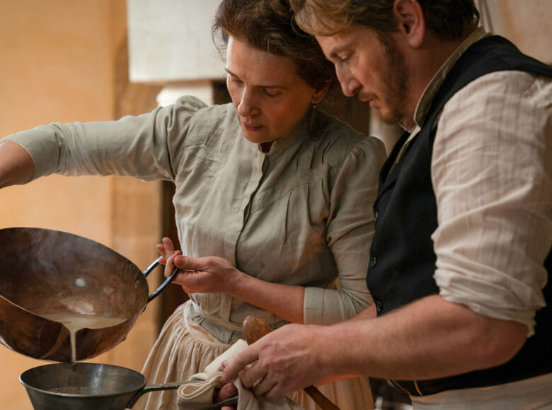 Kolorowa fotografia przedstawia kobietę i mężczyznę w starych, średniowiecznych ubraniach. Kobieta przelewa zawartość garnka przez sito, które trzymane jest przez mężczyznę.