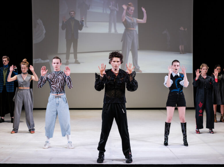 Scena grupowa. 7 osób stoi na białej podłodze, formując ustawieniem literę V. Wszyscy mają ręce uniesione na wysokość twarzy. W tle ekran z powielonym obrazem widocznym na scenie.