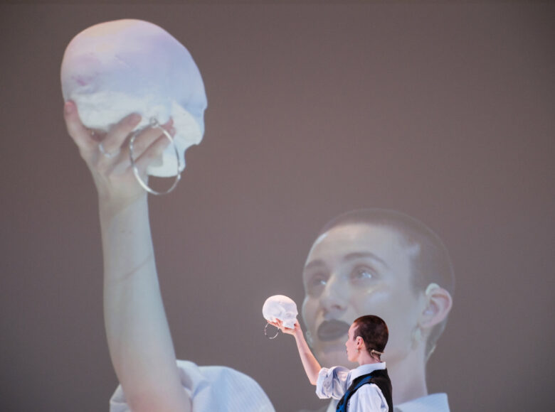 Młoda kobieta trzyma w wyciągniętej nad siebie ręce białą czaszkę, w geście ze monologu Hamleta. Za nią duży ekran powiela obraz widoczny na scenie.