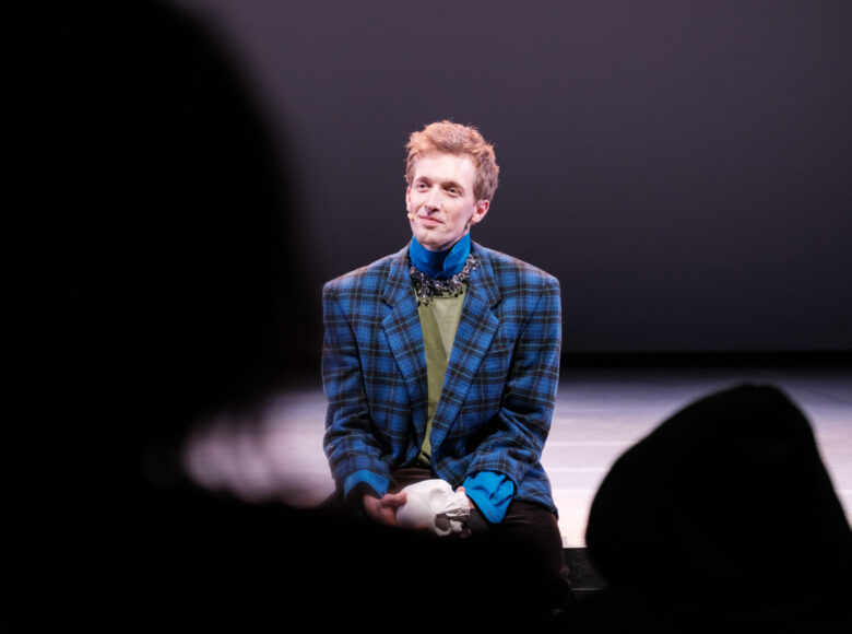Mężczyzna w marynarce w niebieską kratę siedzi na skraju sceny, patrzy przed siebie, częściowo przysłonięty przez głowy widzów.
