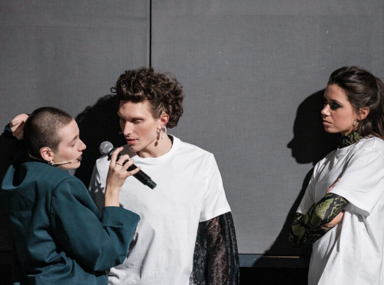 Trzy osoby oparte o szarą ścianę, widoczne od pasa w górę. Dwie osoby po lewej stronie wydają się mówić do mikrofonu, który trzyma jedna z nich. Po prawej młoda kobieta, z rękami złożonymi na piersi, patrzy na pozostałych.