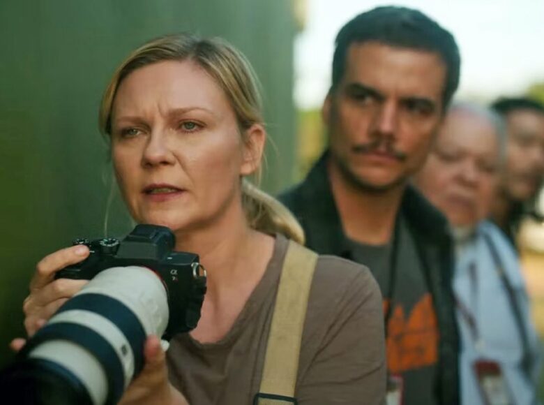 Kadr z filmu. Na zewnątrz. Na pierwszym planie kobieta, trzyma w rękach aparat z długim obiektywem. Za nią stoi mężczyzna i jeszcze dwie osoby, ale każda kolejna osoba jest coraz bardziej nieostra.