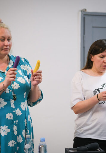 Fotografia przedstawia dwie kobiety. Po lewej, Pani ubrana jest w jasnoniebieską sukienkę w białe kwiaty, ma krótkie, blond włosy. Po prawej kobieta ubrana jest w białą koszulkę z czarnym nadrukiem oraz czarne spodnie. Ma ona ciemne, długie włosy. Obie trzymają małe, kolorowe elementy w dłoniach.