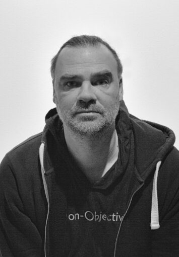 Czarno-biała fotografia przedstawia autoportret mężczyzny o krótkich włosach i brodzie. Znajduje się on w centrum zdjęcia i patrzy prosto w obiektyw. Ubrany jest w rozpinaną bluzę oraz koszulkę.