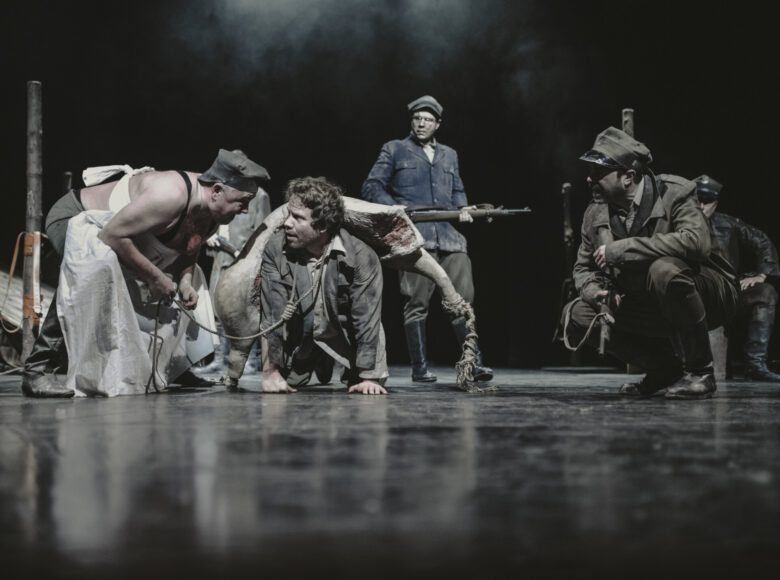 Scena grupowa. Po prawej dwóch mężczyzn klęczy na scenie. Jeden w fartuchu rzeźnika, drugi na plecach ma położony jasny przedmiot. Obok nich po prawej kuca trzeci mężczyzna, w mundurze partyzantów, w tle czwarty mężczyzna stoi i patrzy na akcję z przodu.