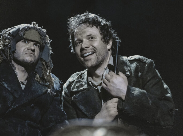 Zbliżenie na dwóch mężczyzn. Jeden trzyma w dłoni karabin, z uśmiechem patrzy przed siebie.