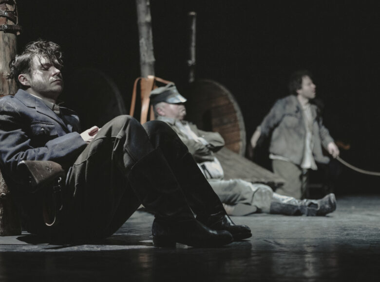 Trzech mężczyzn siedzi na podłodze, plecami są oparci o drewniane pale, opasane skórzanymi pasami.