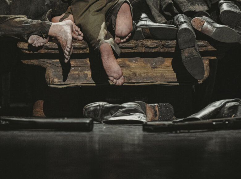 Zbliżenie na nagie stopy mężczyzn, zwisające z krawędzi drewnianego obiektu, na którym leżą. Przed nimi leżą pojedyncze, rozrzucone buty.