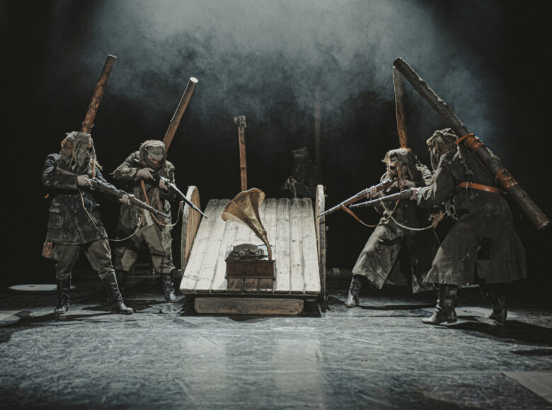 Scena grupowa. Czterech mężczyzn ubranych w poszarpane mundury partyzantów stoi dookoła drewnianej lawety na wysokich kołach. Wszyscy celują karabinami w gramofon z dużą, miedzianą czaszą, stojący na lawecie.