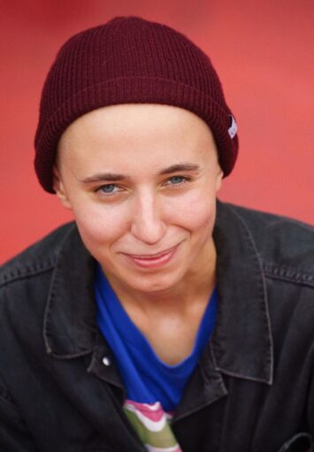 Zdjęcie przedstawia portret osoby patrzącej się prosto w obiektyw z uśmiechem na twarzy. Ubrana jest w czerwoną czapkę typu beanie, niebieską koszulkę oraz czarną, dżinsową kurtkę. W tle jest czerwona ściana.