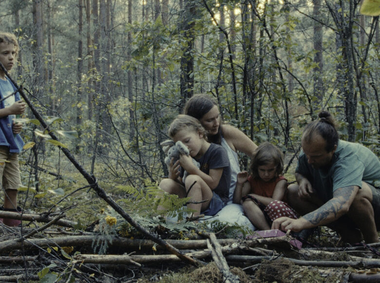 Zdjęcie przedstawia rodzinę w lesie. Od lewej: chłopiec w niebieskiej koszulce i beżowych, krótkich spodenkach, dziecko siedzące matce na kolanach, wtula się w swoją przytulankę, mama w białej sukience siedzi obok córki ubranej na czerwono, a obok córki siedzi ojciec ubrany na zielono. Skrajnie po prawej leży czarny pies z otwartym pyskiem. Cała rodzina znajduje się wśród gałęzi i liści paproci.