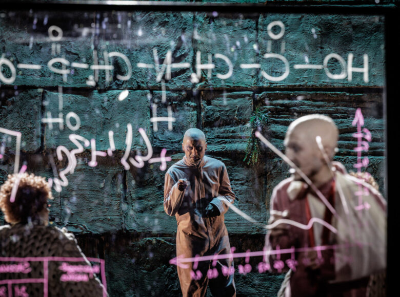 Na zdjęciu trzy osoby, mężczyzna stojący centralnie patrzy prosto w obiektyw. Na pierwszym planie przed nimi przezroczysta tablica, na której są napisane wzory chemiczne.