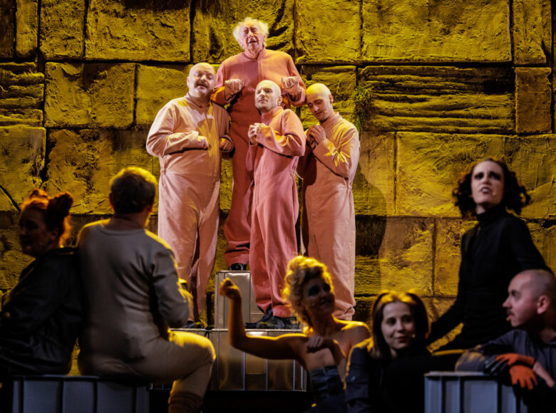 Scena grupowa. Na pierwszym planie osoby w cieniu, siedzące i częściowo niewidoczne. Drugi plan oświetlony, w centrum stoi czterech mężczyzn, ubranych w różowe kombinezony, na tle ściany z dużych bloków kamiennych oświetlonych na żółto.